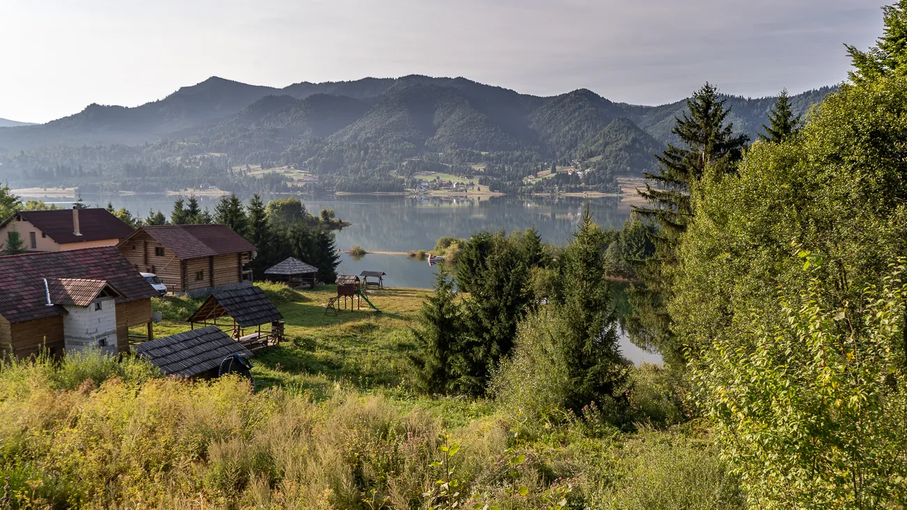 Bergsee mit Holzhütten und bewaldeten Hügeln im Hintergrund