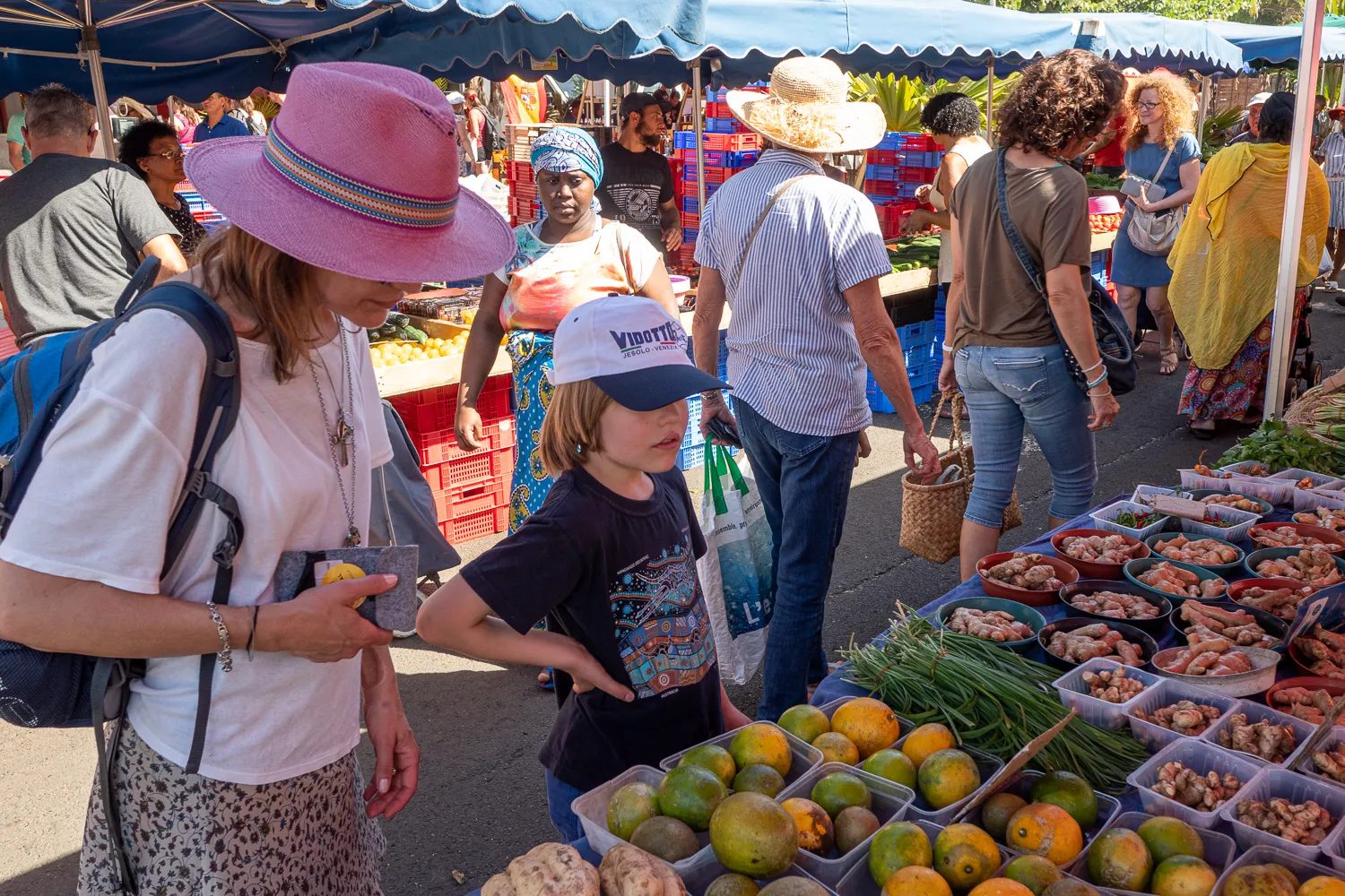 Marktbesucher betrachten Obst und Gemüse unter blauen Markisen.