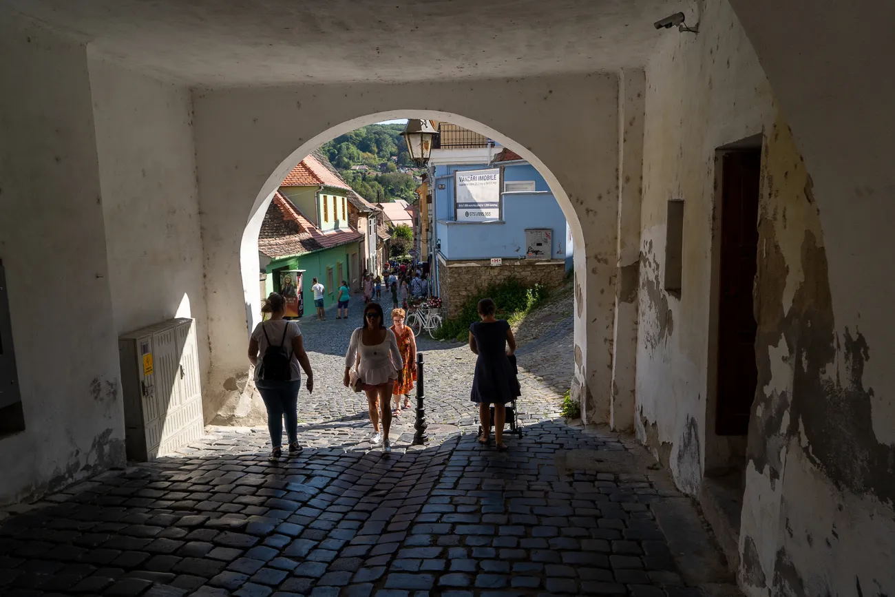 Menschen gehen durch einen steinernen Bogen in eine sonnige Straße mit Häusern.