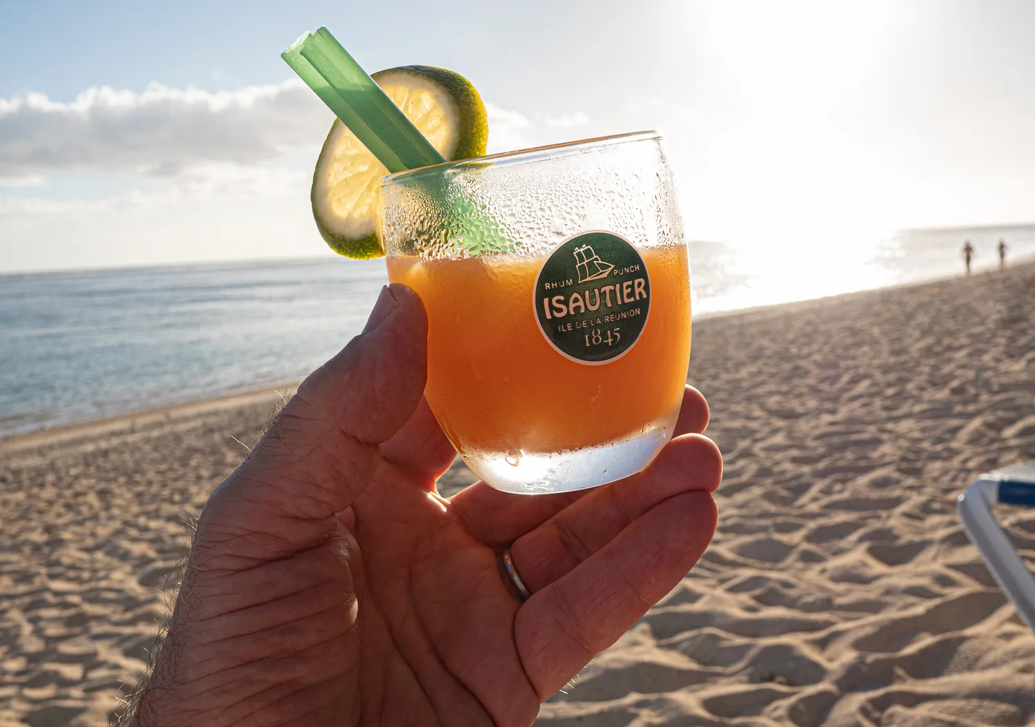 Hand hält Cocktail mit Zitronenscheibe am Strand bei Sonnenuntergang