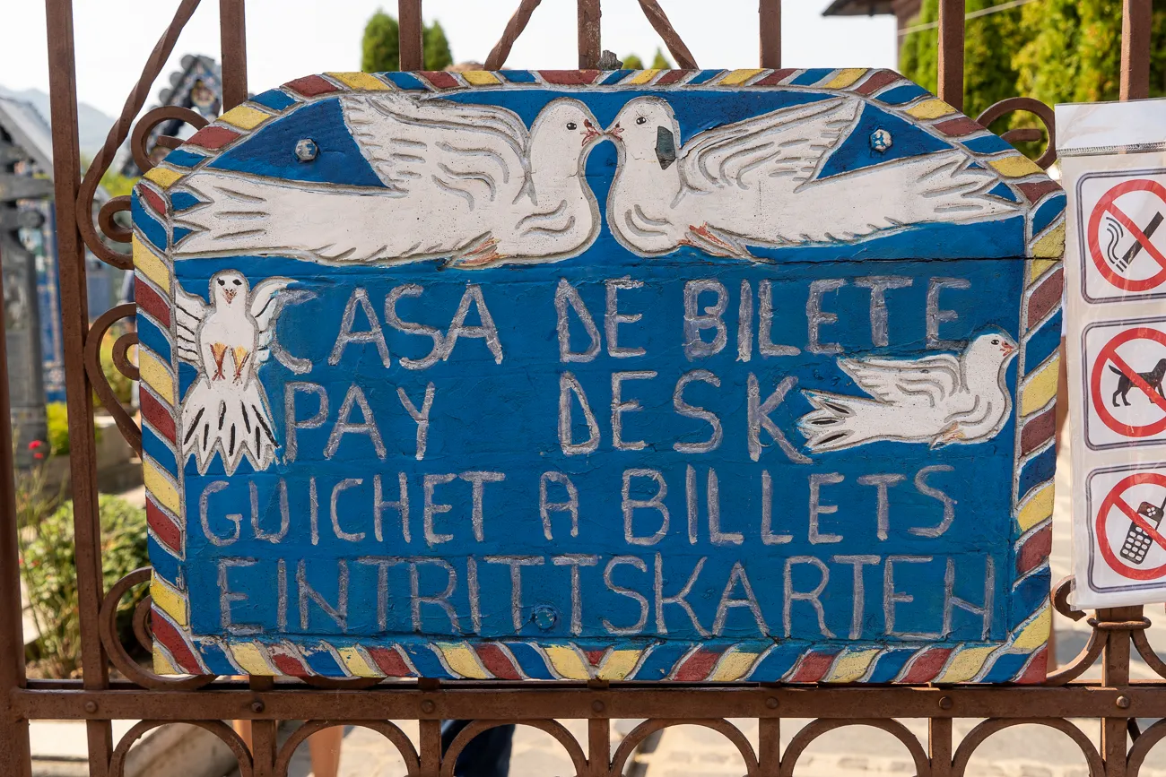Farbiger Holzschild mit Taubenmotiven und mehrsprachigem Eintrittspreis-Schild an einem Metalltor.