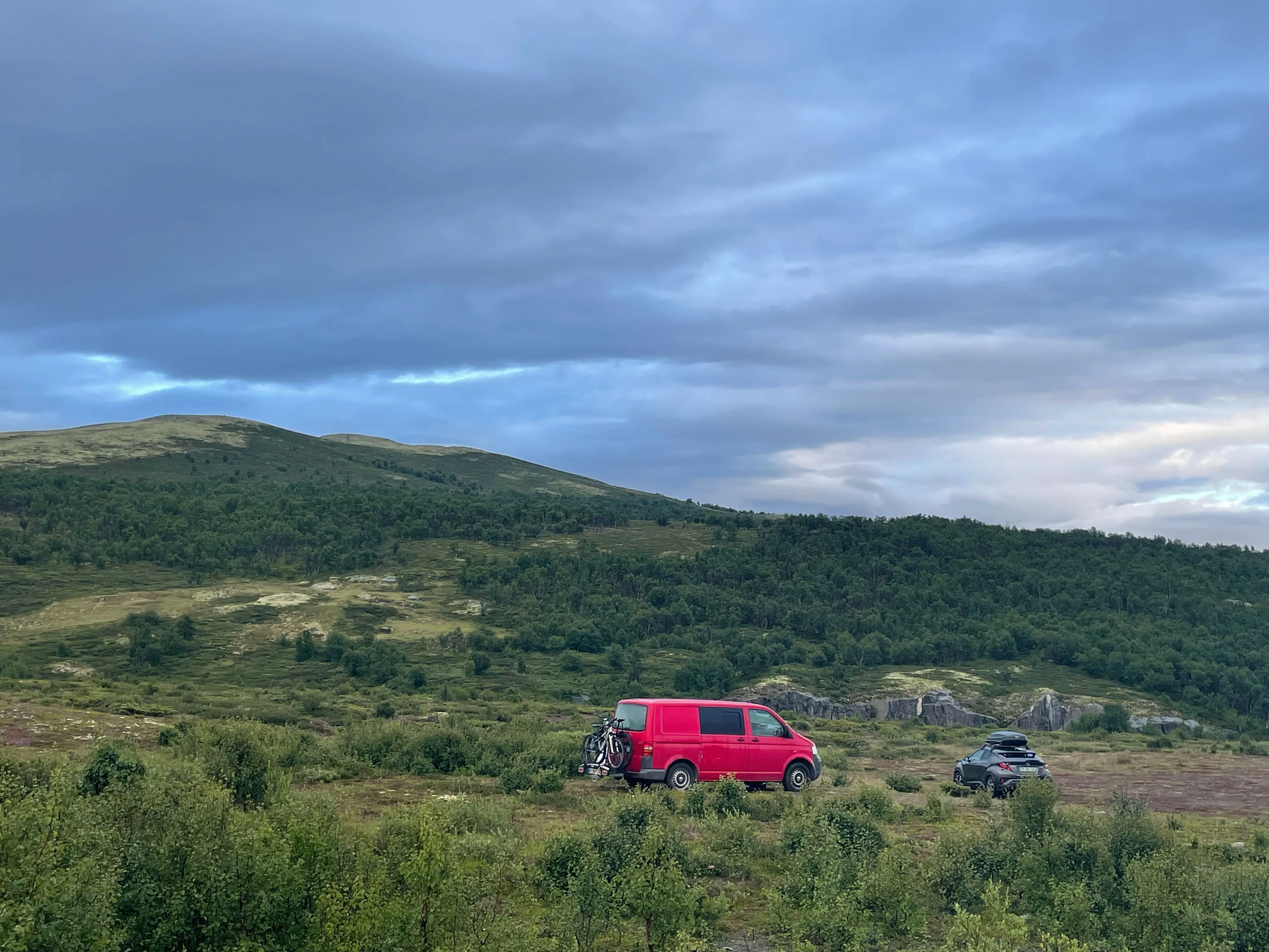 Zwei Fahrzeuge stehen in einer grünen Berglandschaft unter bewölktem Himmel.
