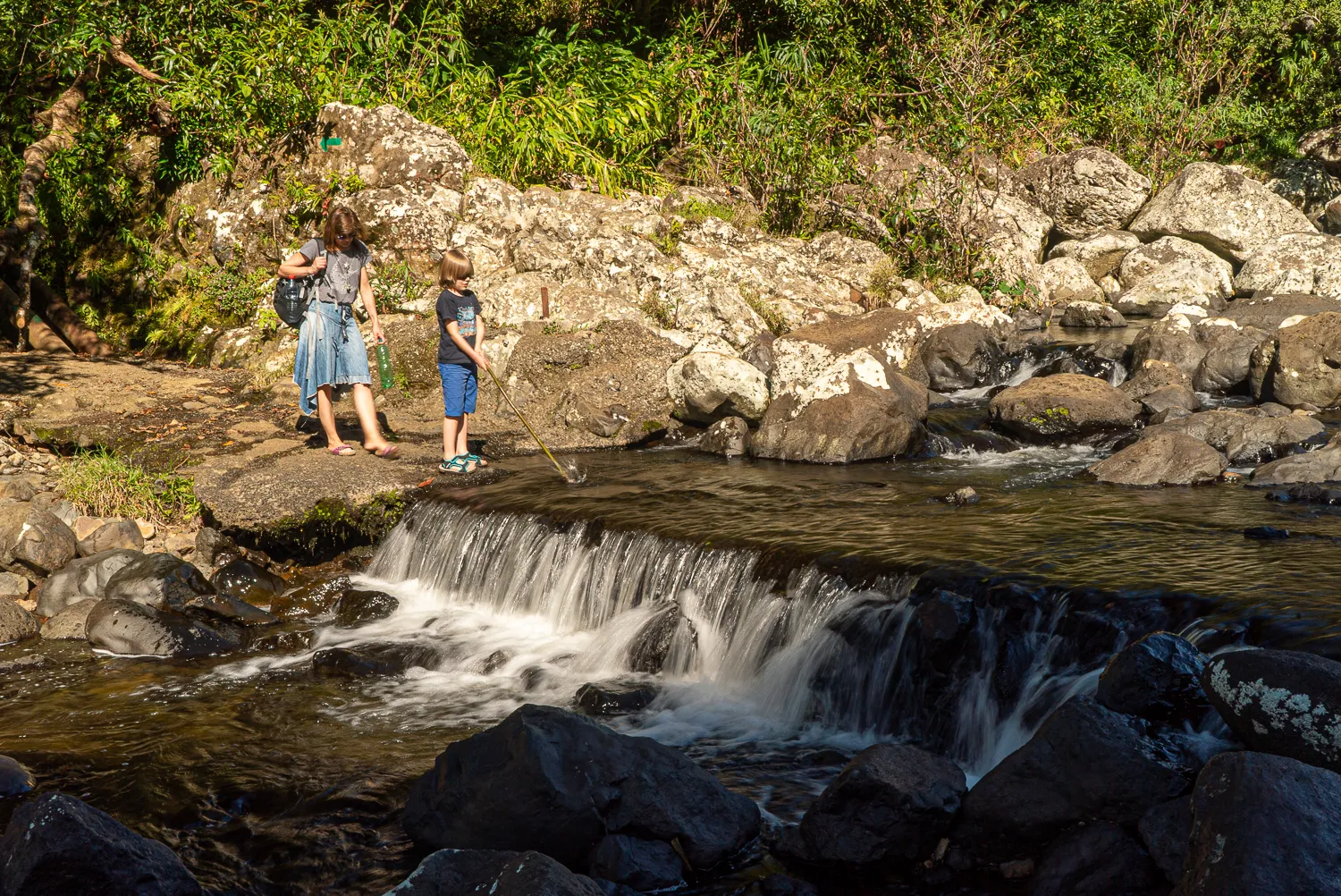 Zwei Kinder stehen am Ufer eines kleinen Wasserfalls in einem steinigen Flussbett.