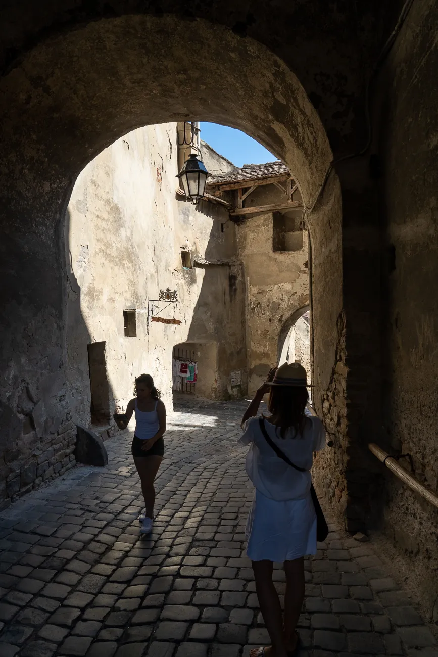 Zwei Personen gehen durch einen steinernen Bogen in eine sonnige Gasse.