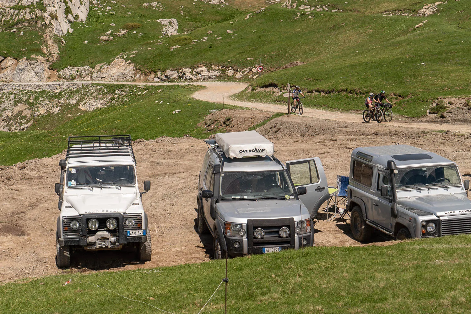 Drei Geländewagen auf einer Kiesbank mit Radfahrern im Hintergrund.