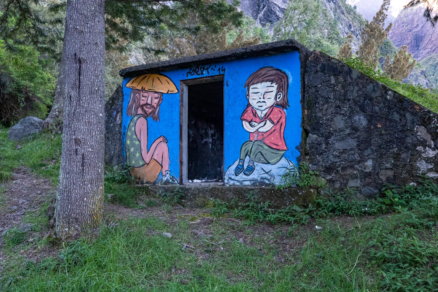 Zwei gemalte Figuren an einer blauen Wand inmitten von Gras und Bäumen