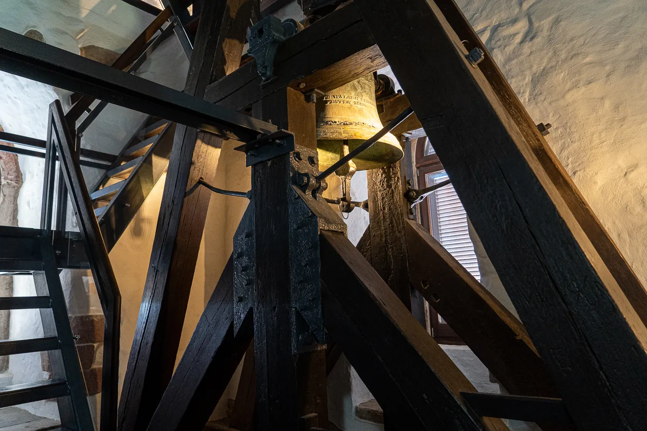 Glocke in hölzernem Rahmen, Wand und Treppe im Hintergrund
