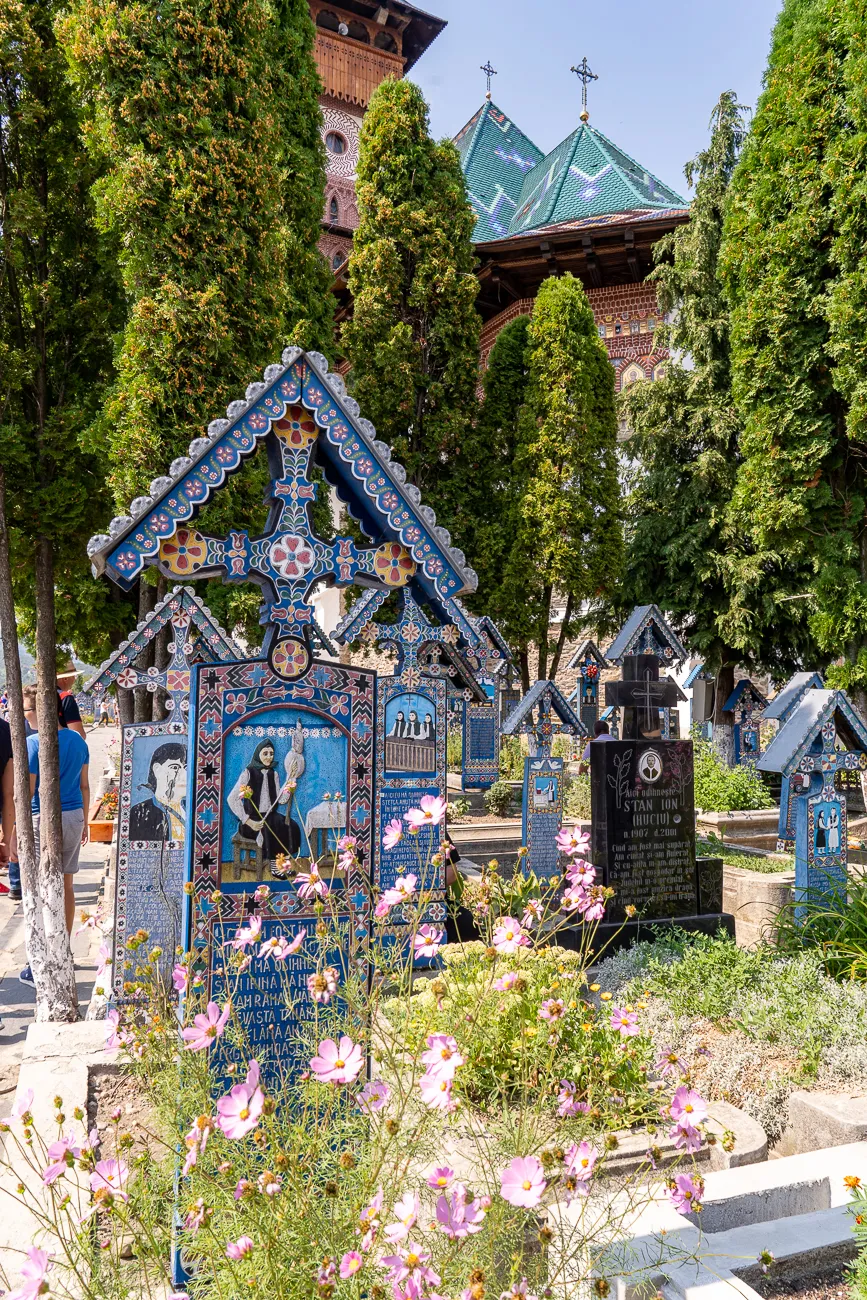 Farbenfrohe Grabkreuze mit religiösen Motiven im Friedhof, umgeben von Blumen und Bäumen.