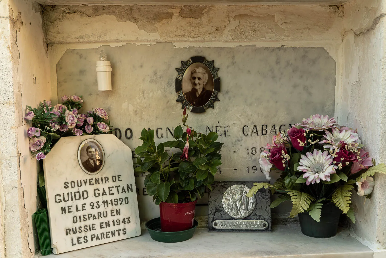Grabstein mit Fotos und Blumen in einem alten Friedhofsbereich.