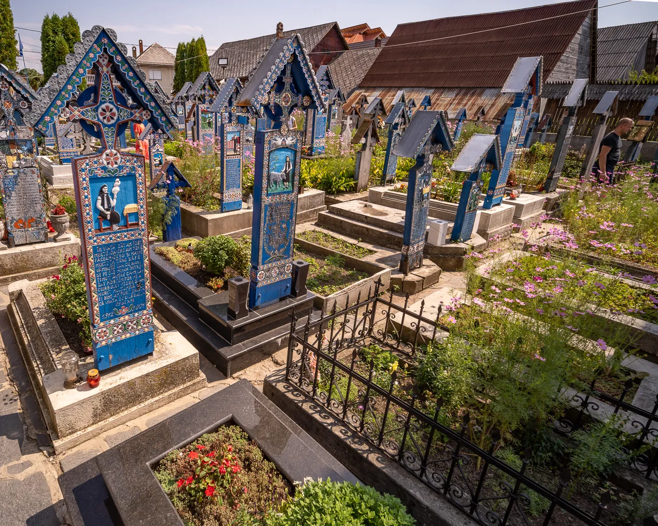 Bunt bemalte Grabsteine in einem Friedhof mit Blumen und Häusern im Hintergrund.