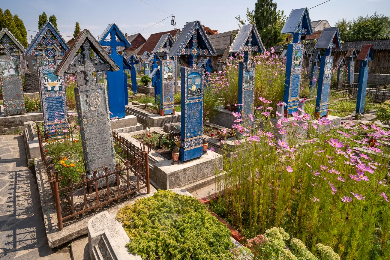 Bunt bemalte Grabsteine in einem Friedhof mit rosa Blumen und grünen Sträuchern.
