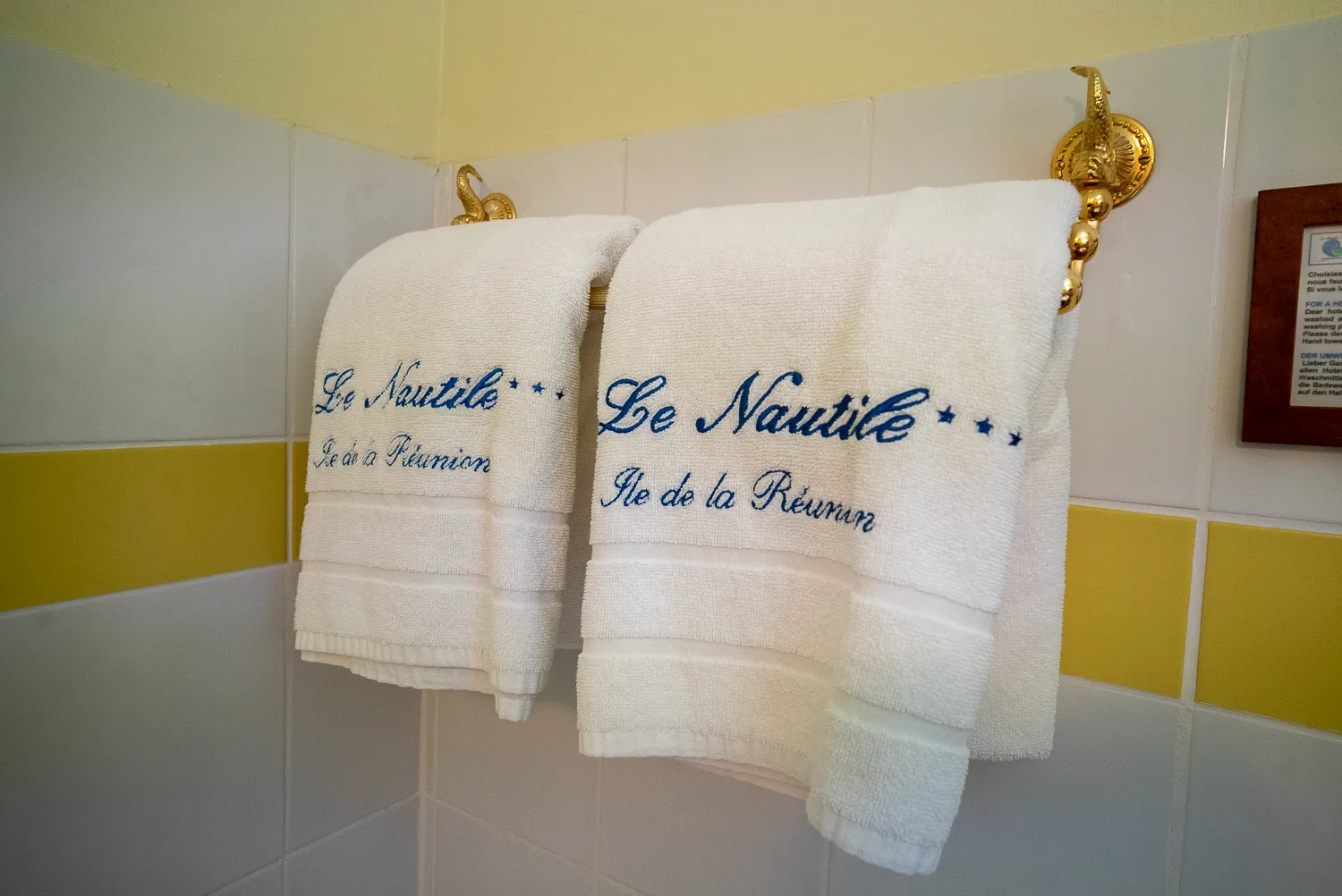 Zwei weiße Handtücher mit Aufschrift Le Nautile, Île de la Réunion, an goldenen Haken in Badezimmer.