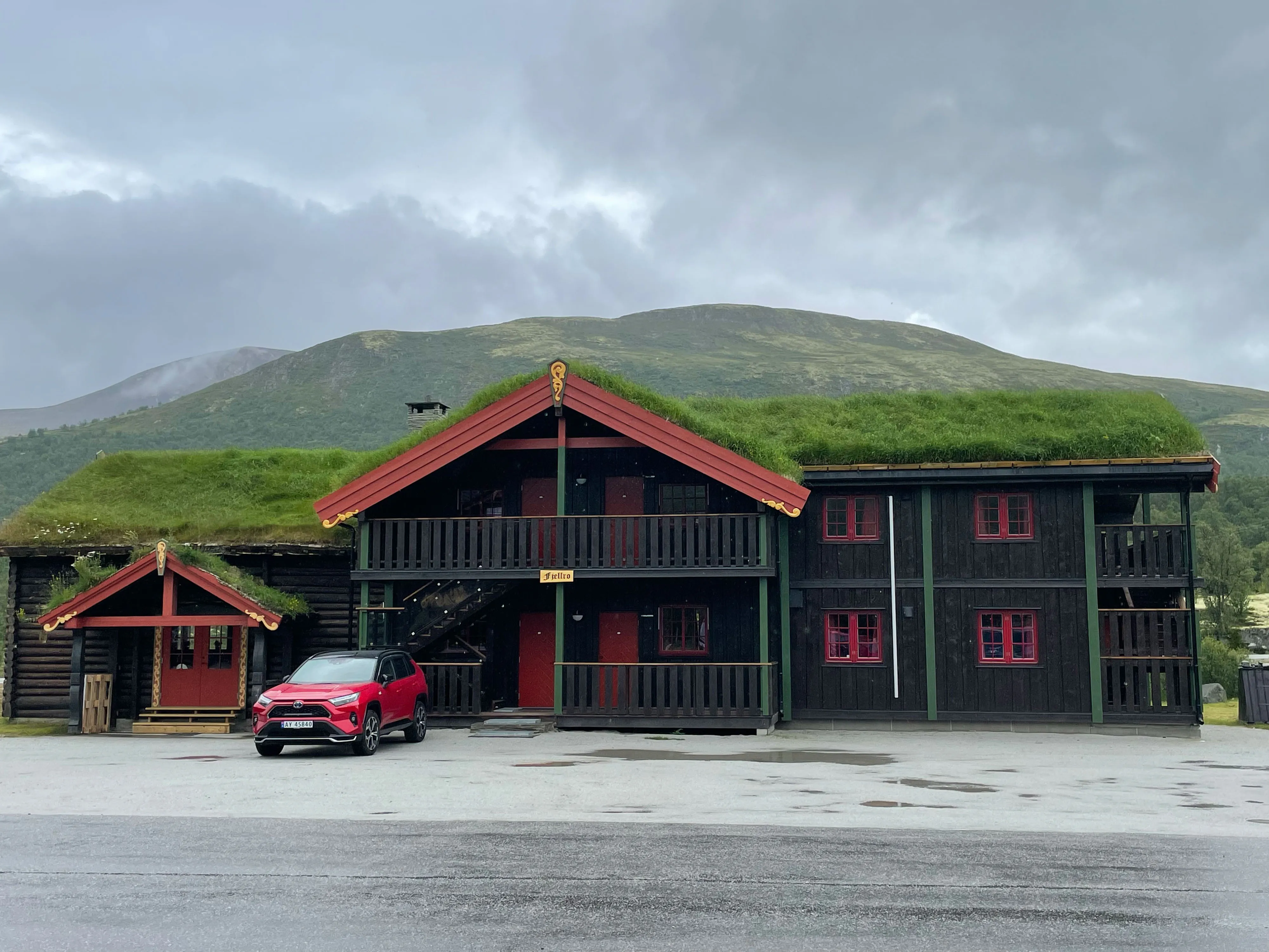 Grüne Dachhaube über dunklem Holzhaus mit rotem Auto vor Berglandschaft