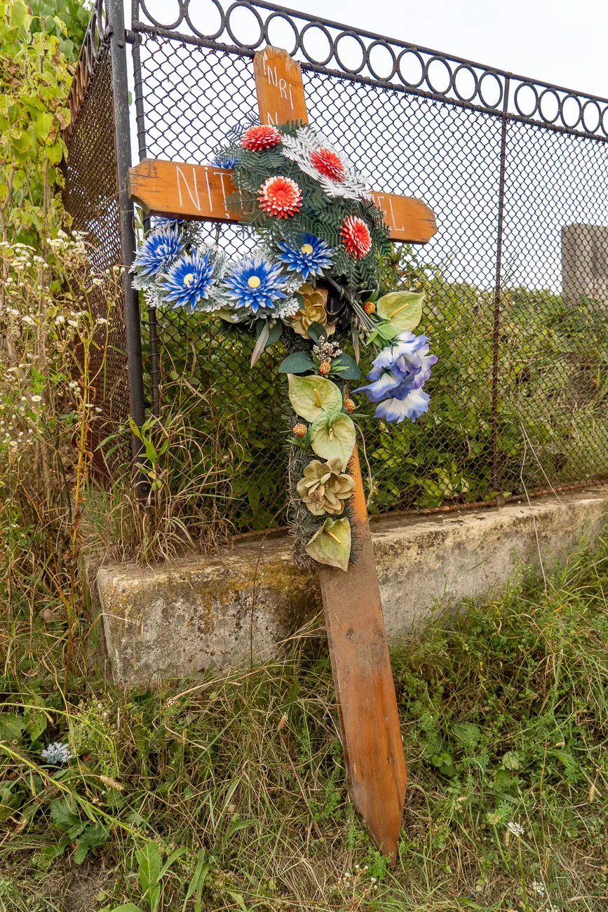 Holzkreuz mit Blumen und Schriftzug vor Zaun und Steinmauer