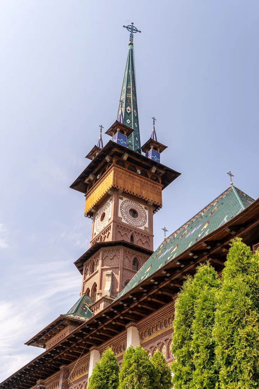 Hoch aufragender Holzturm mit grünem Zeltdach und Kreuz, umgeben von Nadelbäumen.