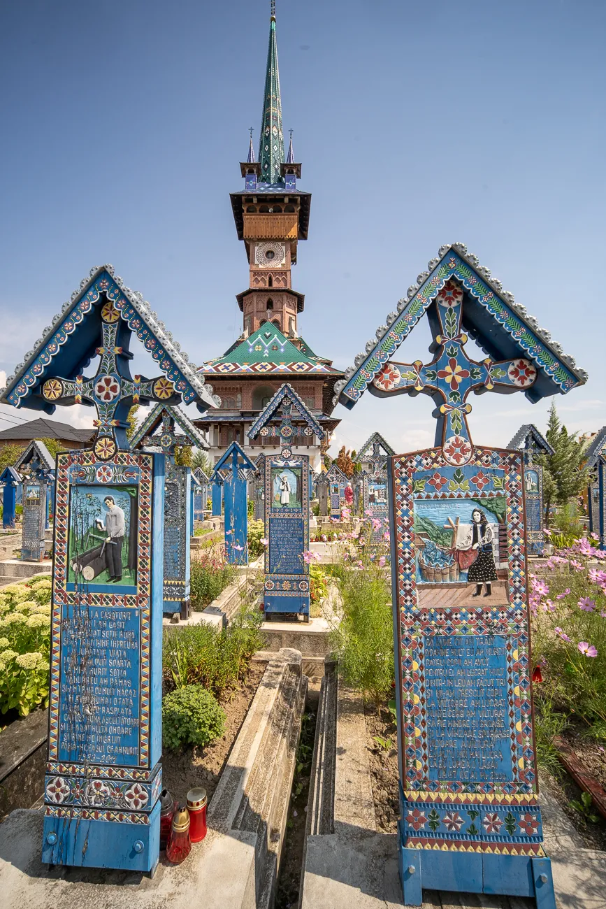 Farbige Holzkreuze im Friedhof mit bemalten Szenen und Inschriften, im Hintergrund ein Kirchturm.