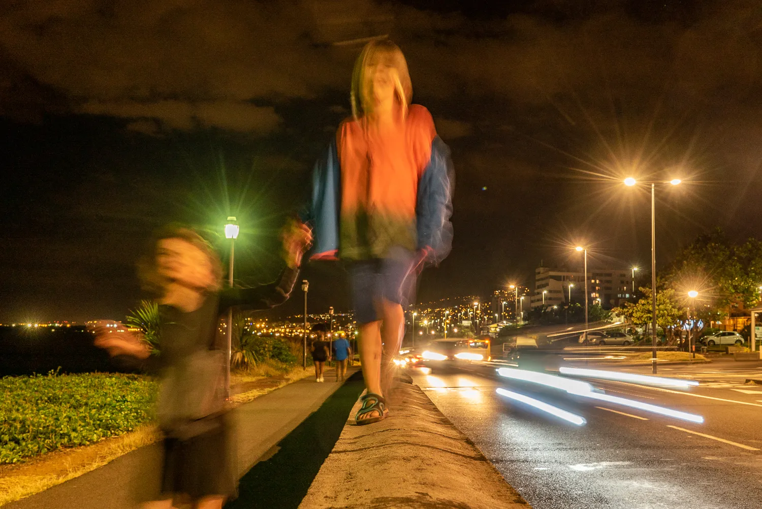 Nachtansicht mit Lichtspuren, Personen auf Gehweg, Straßenbeleuchtung und Stadtlichter im Hintergrund