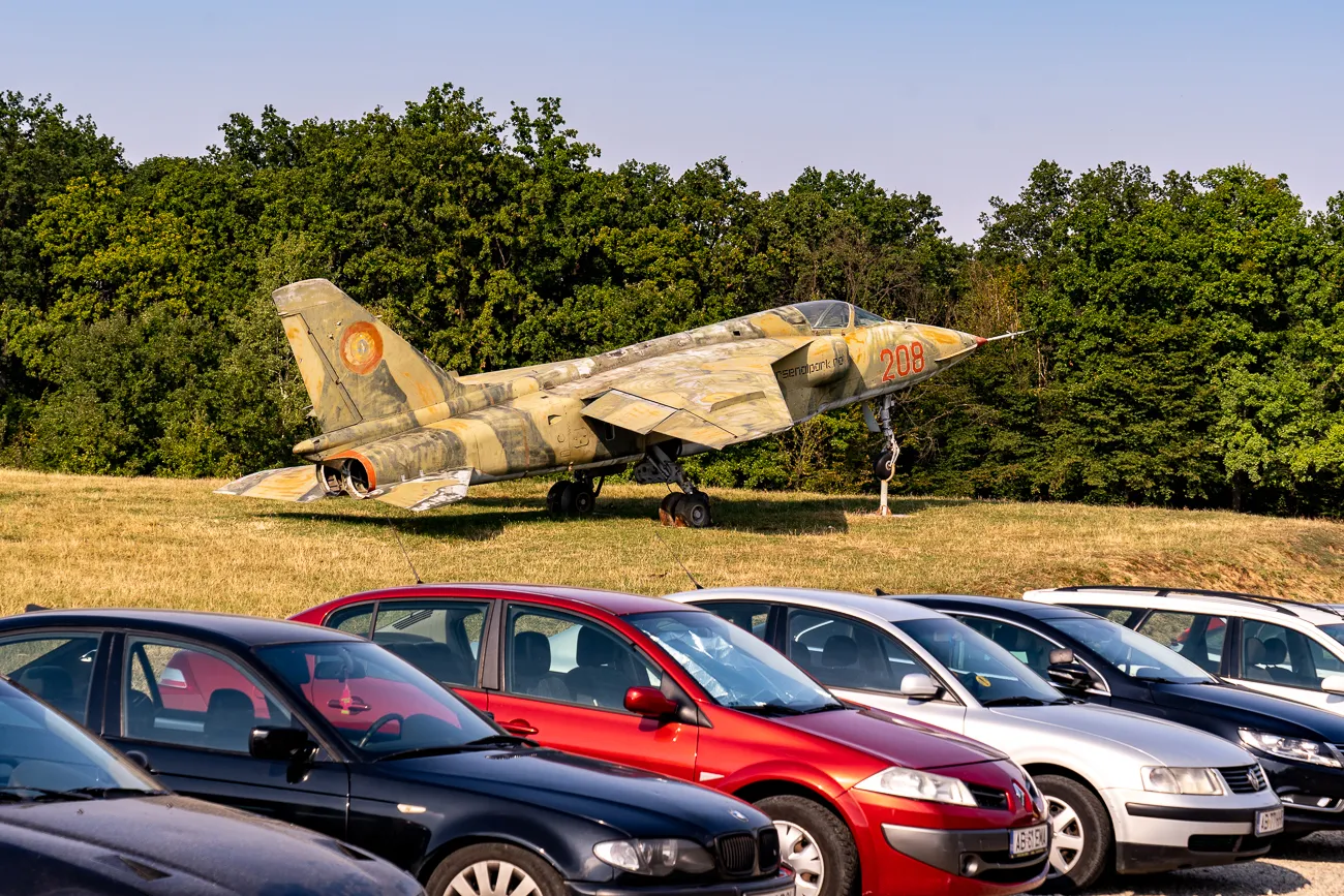 Verwitterter Militärjet auf einer Wiese hinter parkenden Autos.