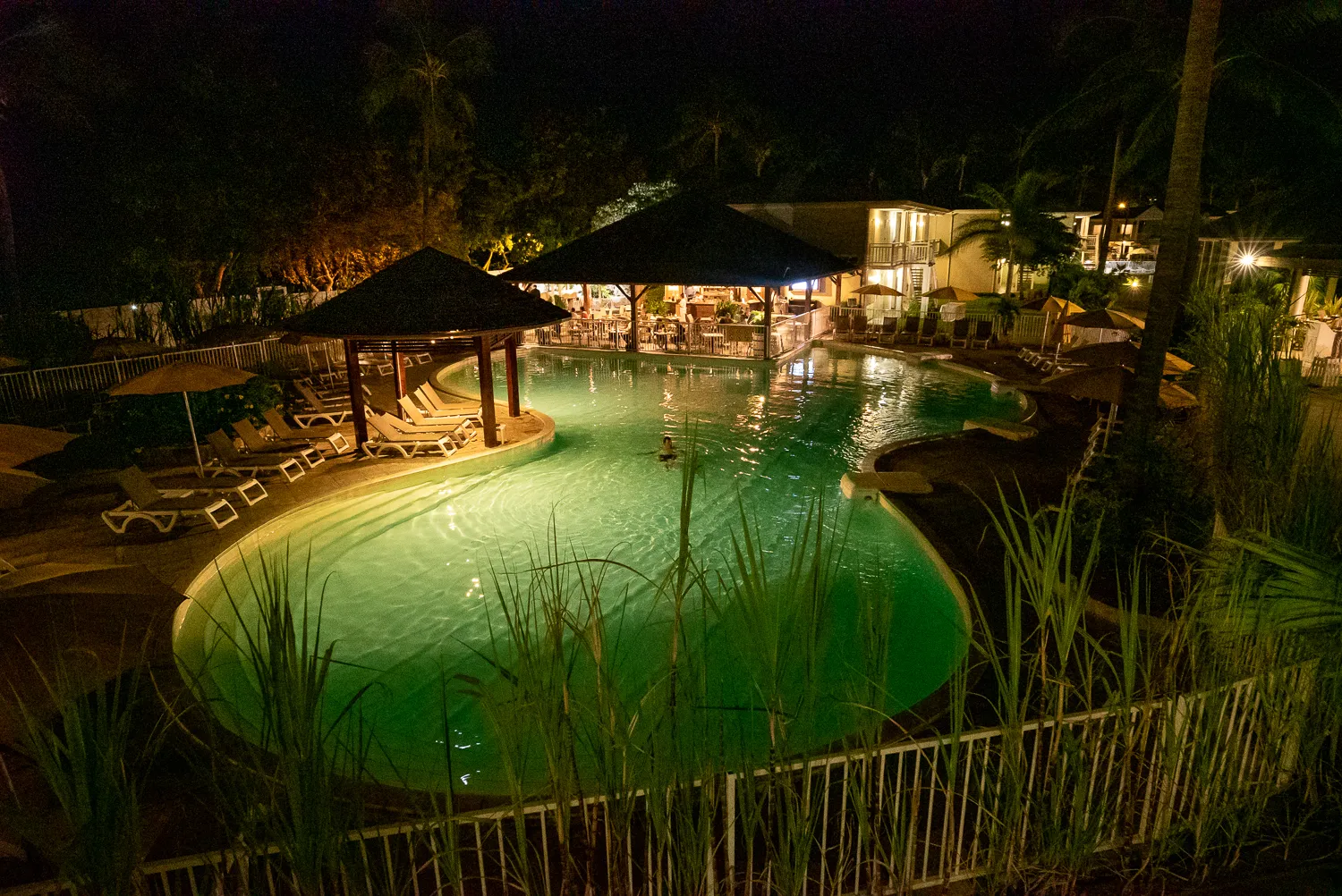 Nachtansicht eines beleuchteten Pools mit Liegestühlen und Palmen