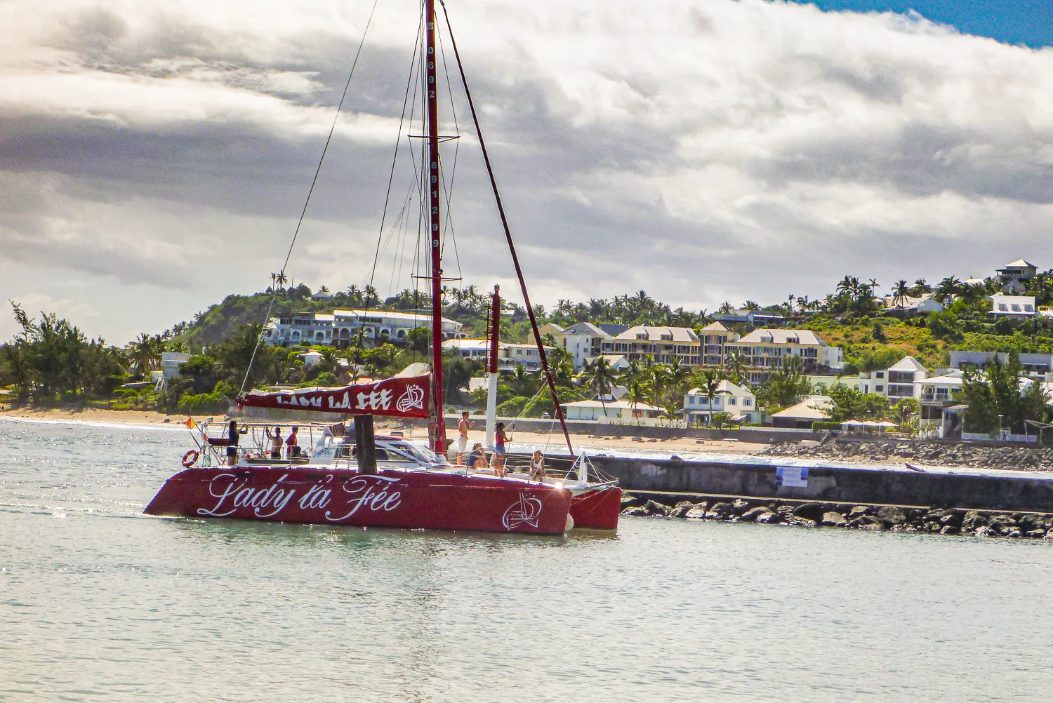 Rotes Katamaranboot mit Schriftzug auf dem Wasser vor Strand und Häusern