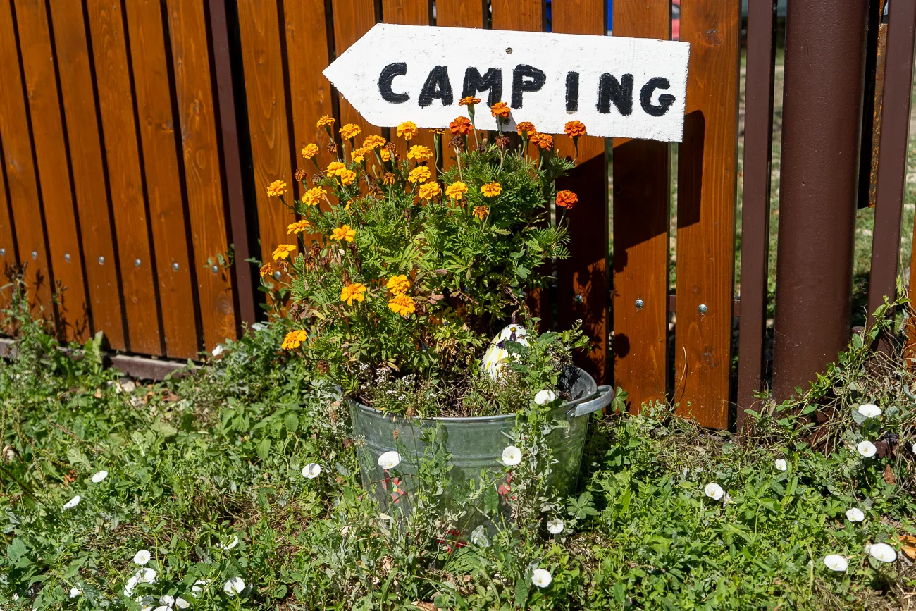 Schild mit Camping-Schriftzug vor Holzzaun und Blumenkübel