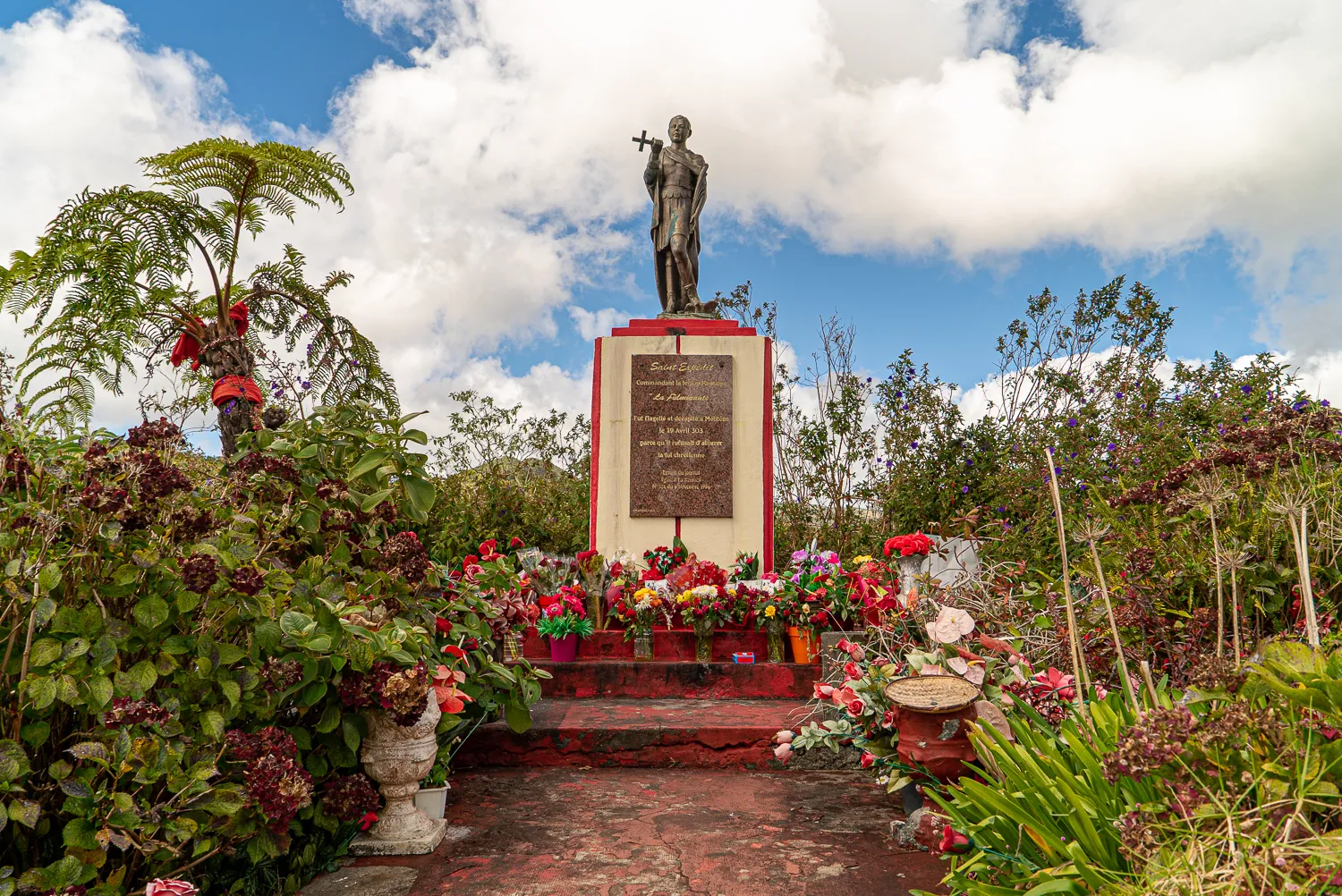 Statue auf Säule mit Blumen, umgeben von Pflanzen und Wolkenhimmel