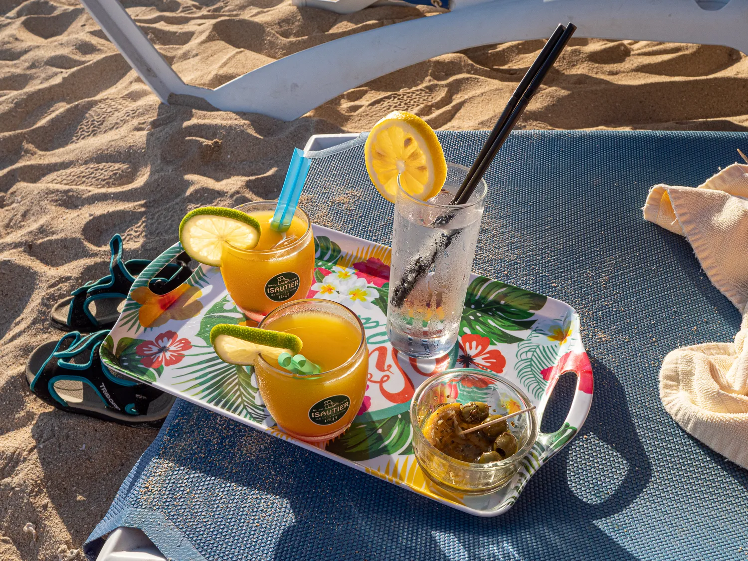 Getränke und Snacks auf einem Strandtisch mit Sand und Liegestuhl