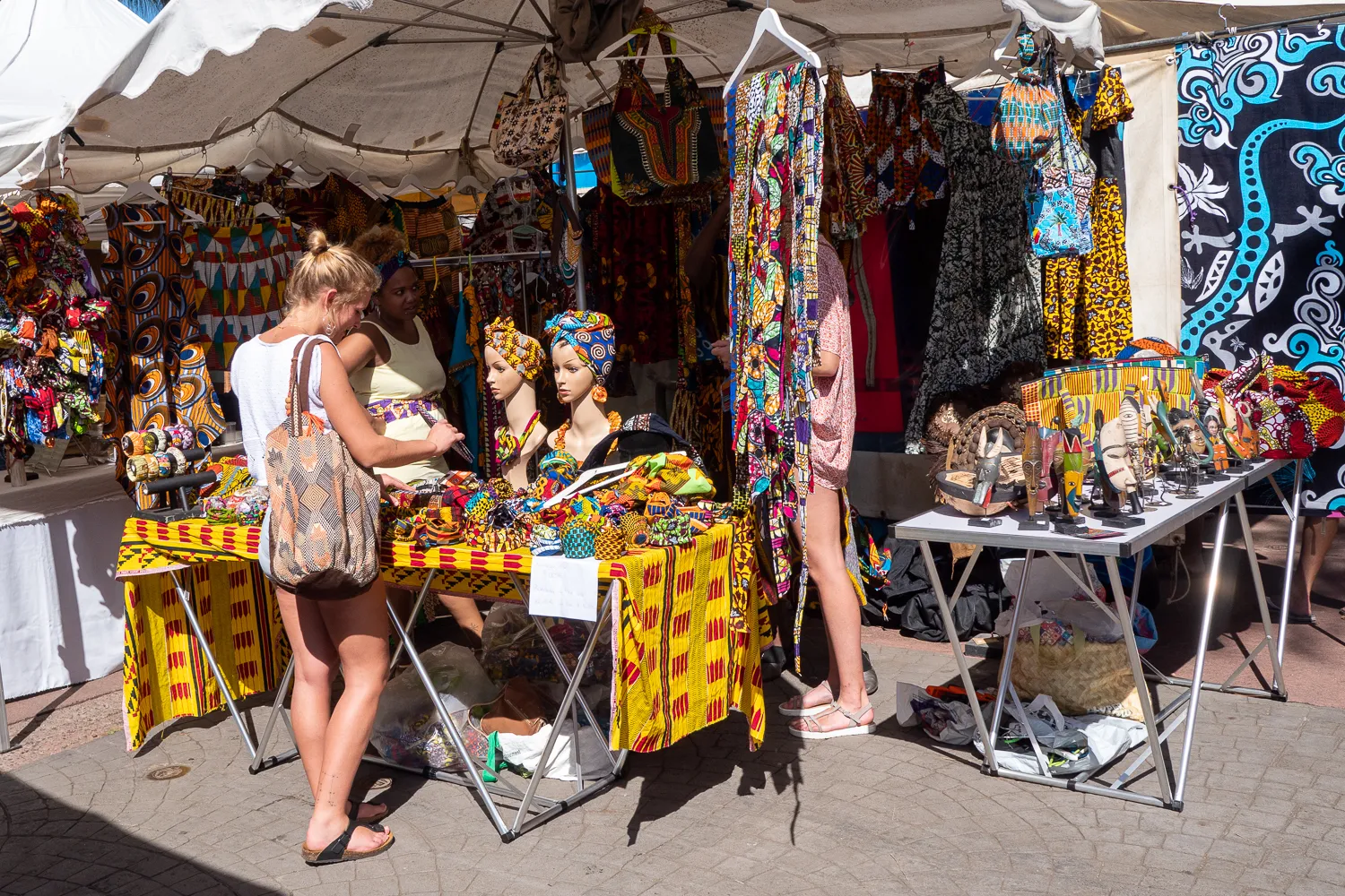 Frauen betrachten farbenfrohe Textilien und Souvenirs auf einem Marktstand.