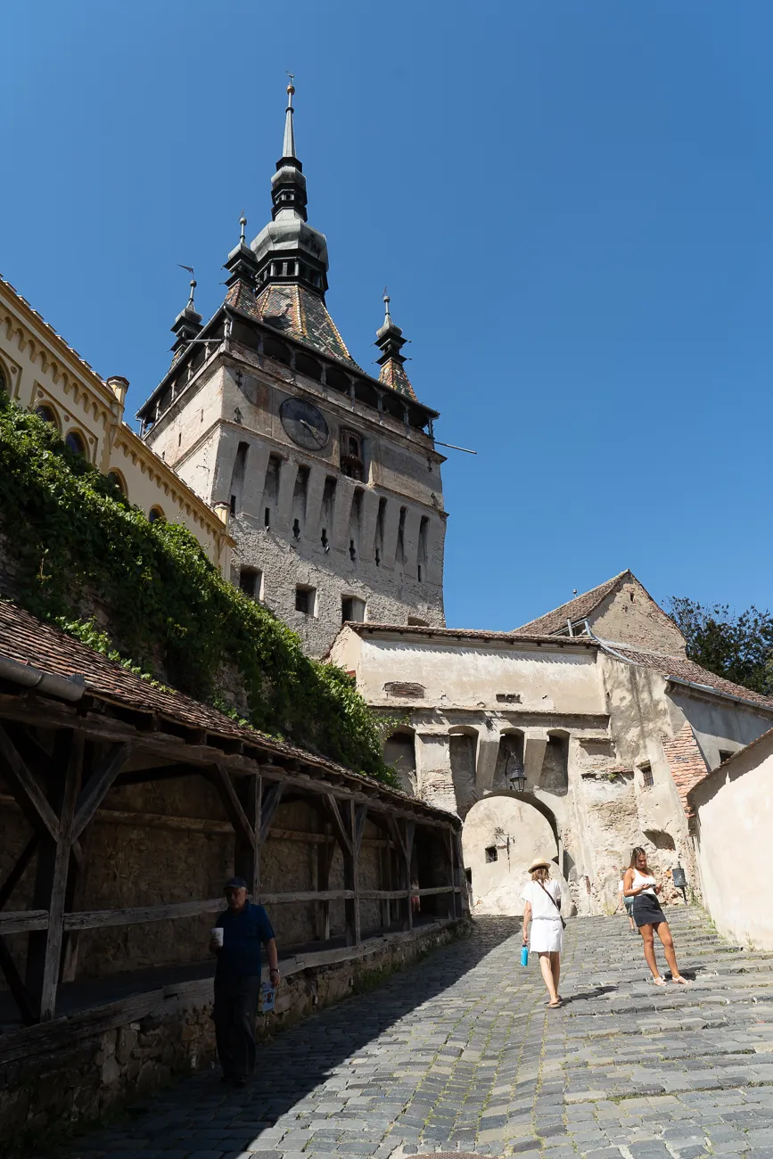 Hoch aufragender Turm mit Uhr und Zinnen unter blauem Himmel, steinerner Weg mit Passanten.