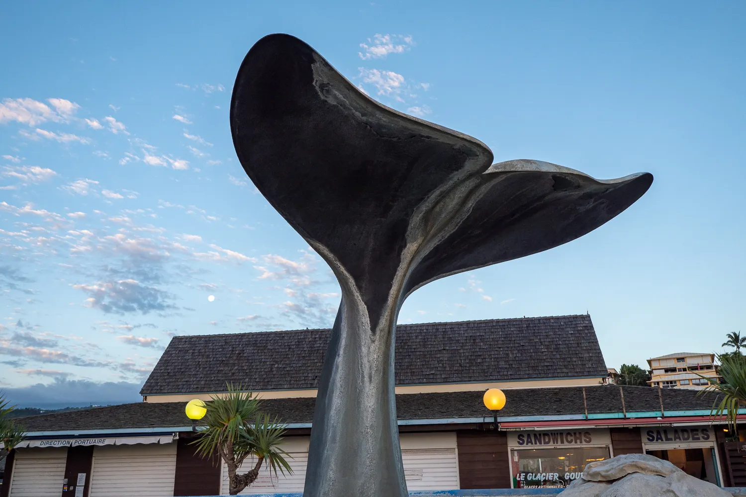 Metallskulptur von Walzflosse vor Strandhütten mit Palmen und blauem Himmel.