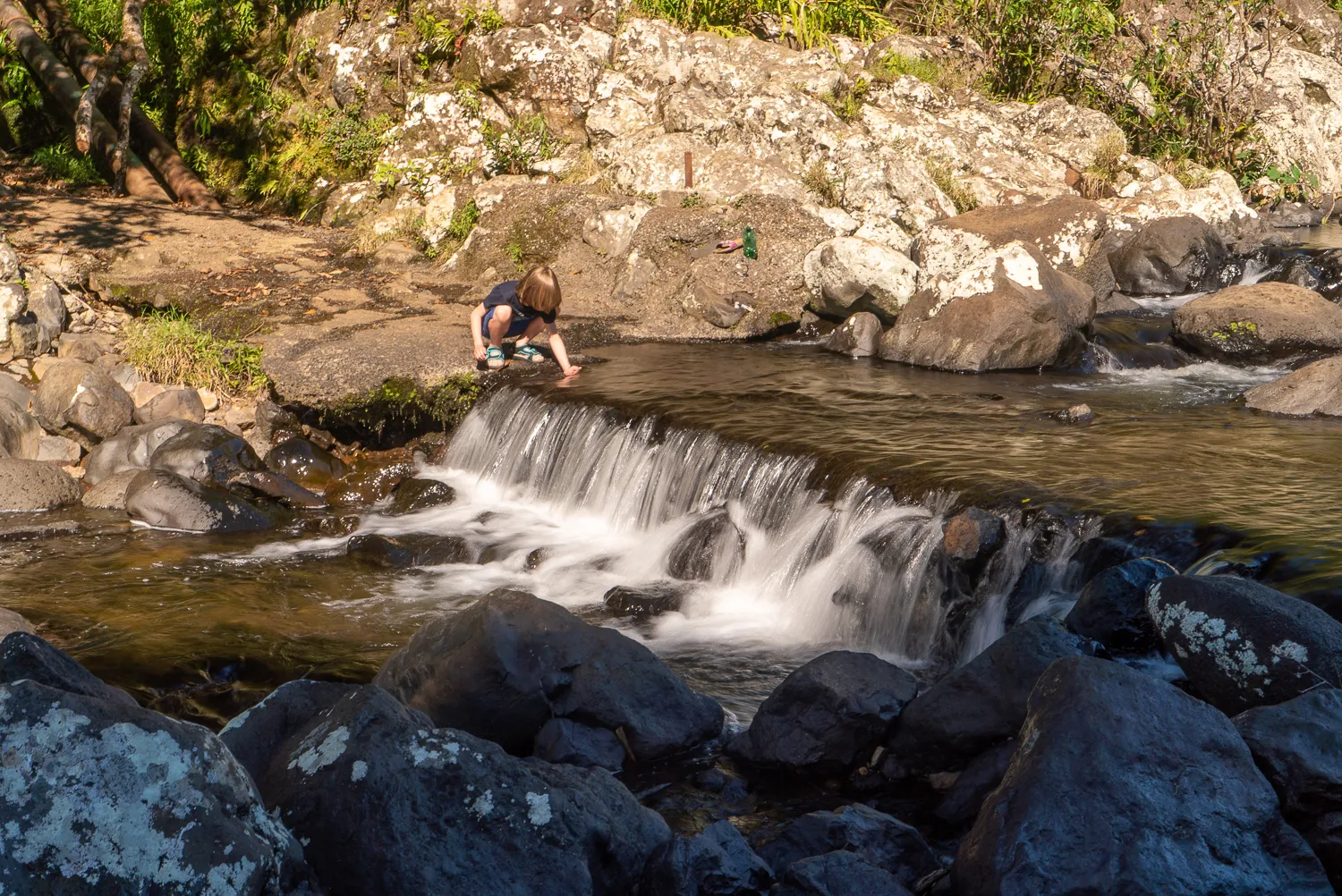 Mensch kniet am Wasserfallrand, fließendes Wasser über Felsen.