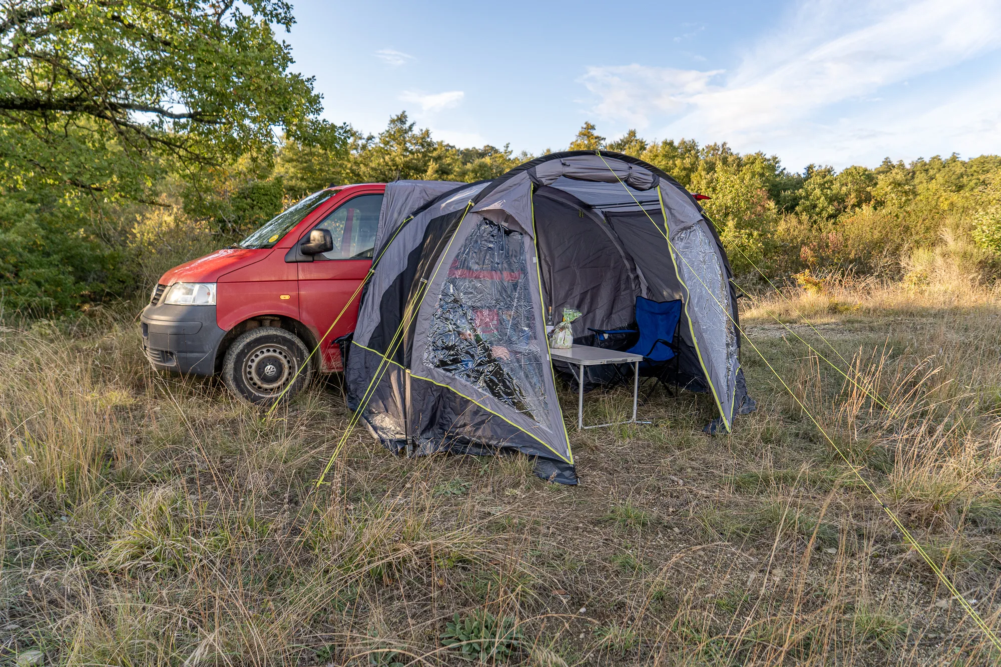Rot lackierter Kastenwagen neben einem Zelt im Gras unter Bäumen.